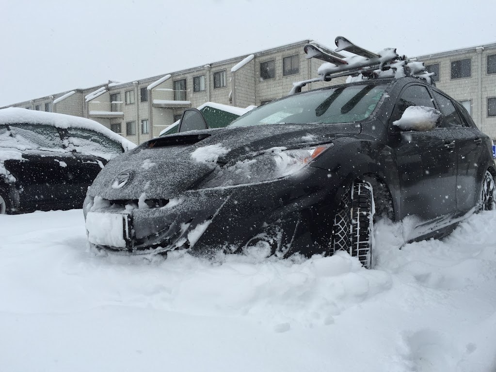 A dark hatchback with skis on its roof rack sits in deep snow in a Snowshoe parking lot, snow still falling, a condo building behind it.